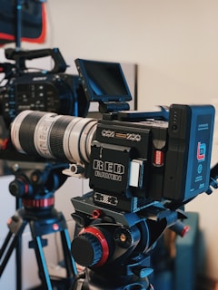 A professional video camera setup is mounted on a tripod. The camera features a large lens and a monitor screen, with various dials and buttons visible on the equipment. Another camera and tripod can be seen in the background.