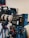 A professional video camera setup is mounted on a tripod. The camera features a large lens and a monitor screen, with various dials and buttons visible on the equipment. Another camera and tripod can be seen in the background.
