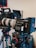 A professional video camera setup is mounted on a tripod. The camera features a large lens and a monitor screen, with various dials and buttons visible on the equipment. Another camera and tripod can be seen in the background.