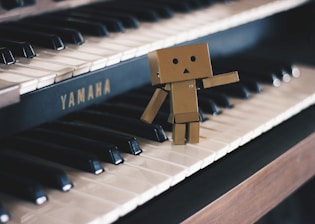 A small cardboard robot figure stands on the keys of a Yamaha piano, appearing to wave or gesture with one arm. The piano's black and white keys create a strong contrast with the robot's brown color, and the lighting adds a soft tone to the scene.