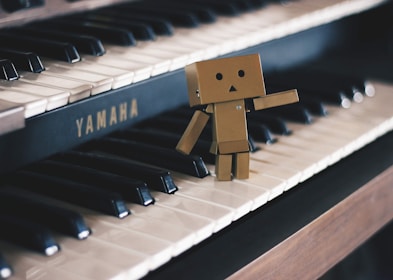 A small cardboard robot figure stands on the keys of a Yamaha piano, appearing to wave or gesture with one arm. The piano's black and white keys create a strong contrast with the robot's brown color, and the lighting adds a soft tone to the scene.