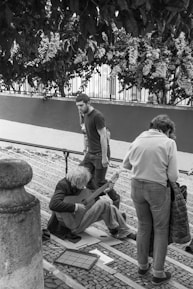 A street musician sits on cobblestone pavement playing a guitar. A person in a white jacket is standing nearby, seemingly observing or interacting with the musician. Another person walks past, looking down at the musician. The scene is set in an outdoor environment with leafy foliage and a decorative fence in the background.