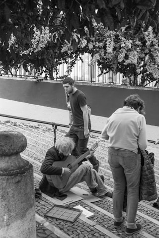 A street musician sits on cobblestone pavement playing a guitar. A person in a white jacket is standing nearby, seemingly observing or interacting with the musician. Another person walks past, looking down at the musician. The scene is set in an outdoor environment with leafy foliage and a decorative fence in the background.