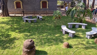 A lush green lawn with two circular concrete benches arranged in a garden setting. A stone building with two closed windows and a wooden fence is visible in the background. A few trees and shrubs are scattered throughout the area, and a group of people is walking in the distance.