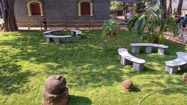 A lush green lawn with two circular concrete benches arranged in a garden setting. A stone building with two closed windows and a wooden fence is visible in the background. A few trees and shrubs are scattered throughout the area, and a group of people is walking in the distance.