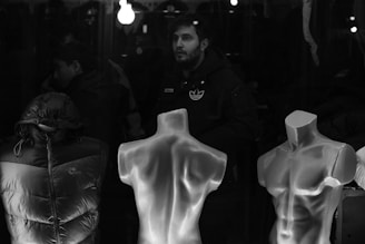 A black and white image depicting a storefront window display. Two mannequin torsos are in the foreground; one is covered by a puffer jacket and the other is bare. In the background, a person in a dark jacket with a visible logo stands under dim lighting, with reflections and hanging lights visible.