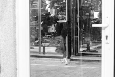 A monochrome image of a storefront display. The reflection on the glass door shows stacked clothing items inside a retail store. A mannequin dressed in trousers stands in the doorway, and various other clothing items are visible behind it. The scene captures a typical clothing shop setup with a transparent view of the store interior from outside.