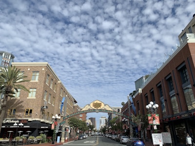 A city street scene with a large decorative archway reading 'Historic Heart of San Diego' spanning across the road. The street is lined with multi-story brick buildings, featuring shops and restaurants, including a place called Mickey's. The sky is filled with a pattern of wavy, thin clouds. Several cars are parked along the sides of the street, and palm trees and decorative street lamps add to the urban setting.
