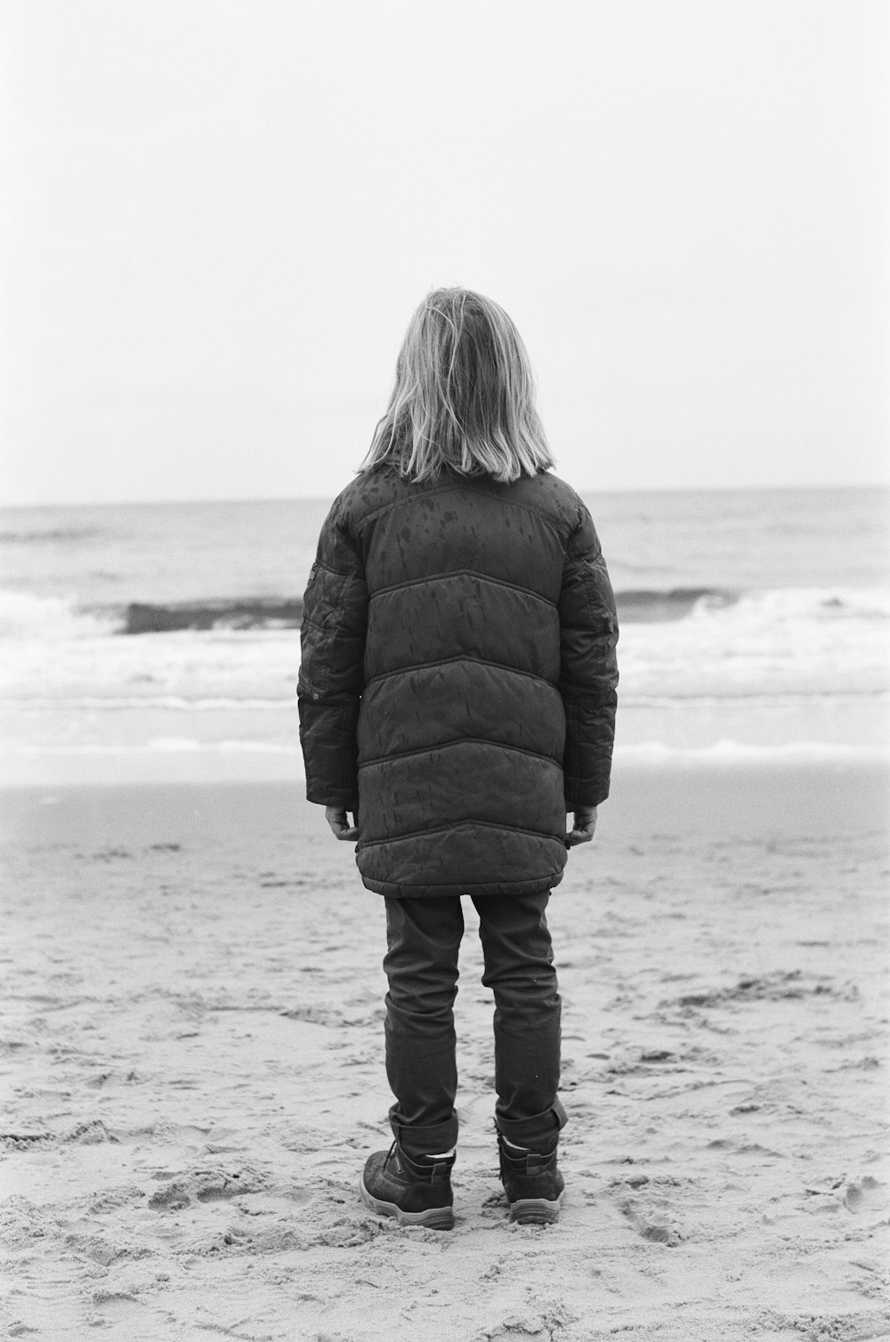 grayscale photography of person wearing down jacket standing on shore during daytime