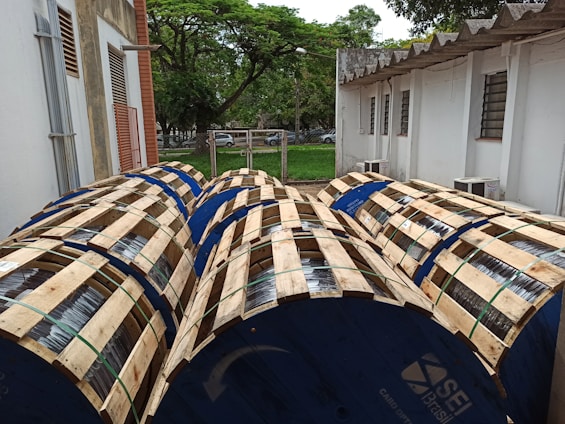 Several large wooden reels wrapped in metal wiring are placed outdoors in a yard beside a building. The reels are blue, bounded by wooden slats, and are lined up next to each other. Behind them is a white building with a corrugated roof and louvered windows. A lush green lawn and trees can be seen in the background.