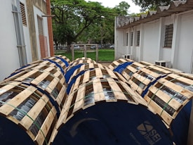 Several large wooden reels wrapped in metal wiring are placed outdoors in a yard beside a building. The reels are blue, bounded by wooden slats, and are lined up next to each other. Behind them is a white building with a corrugated roof and louvered windows. A lush green lawn and trees can be seen in the background.