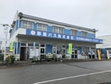 A two-story blue and white building with a large sign in Japanese characters on the facade. The building features multiple windows and doors, with some people standing near the entrance. Vending machines are visible on one side, and various banners hang around the entrance area. Potted plants and a railing line the front of the building.