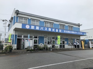 A two-story blue and white building with a large sign in Japanese characters on the facade. The building features multiple windows and doors, with some people standing near the entrance. Vending machines are visible on one side, and various banners hang around the entrance area. Potted plants and a railing line the front of the building.