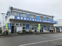 A two-story blue and white building with a large sign in Japanese characters on the facade. The building features multiple windows and doors, with some people standing near the entrance. Vending machines are visible on one side, and various banners hang around the entrance area. Potted plants and a railing line the front of the building.