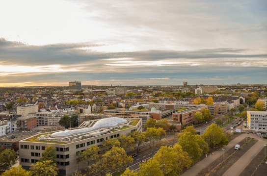 Aerial view of a cityscape during sunset, with numerous buildings, streets, and dense greenery interspersed throughout the area. The sky is partly cloudy with the sun creating a warm glow over the horizon. A unique building with an interesting roof design is prominently visible in the foreground.