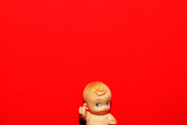 A small, vintage-style plastic baby doll with blond hair and blue eyes is positioned against a vivid red background. The doll is located at the bottom of the image, gazing upward with its right hand raised.