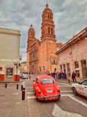 red Volkswagen Beetle passing by brown building during daytime