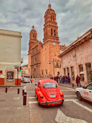 red Volkswagen Beetle passing by brown building during daytime