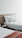 A neatly made bed with white pillows and sheets, alongside a wooden headboard. Next to it, a wooden nightstand holds a telephone and some papers. A folded red and orange blanket lies at the foot of the bed, adding a splash of color to the otherwise minimalistic and bright room.