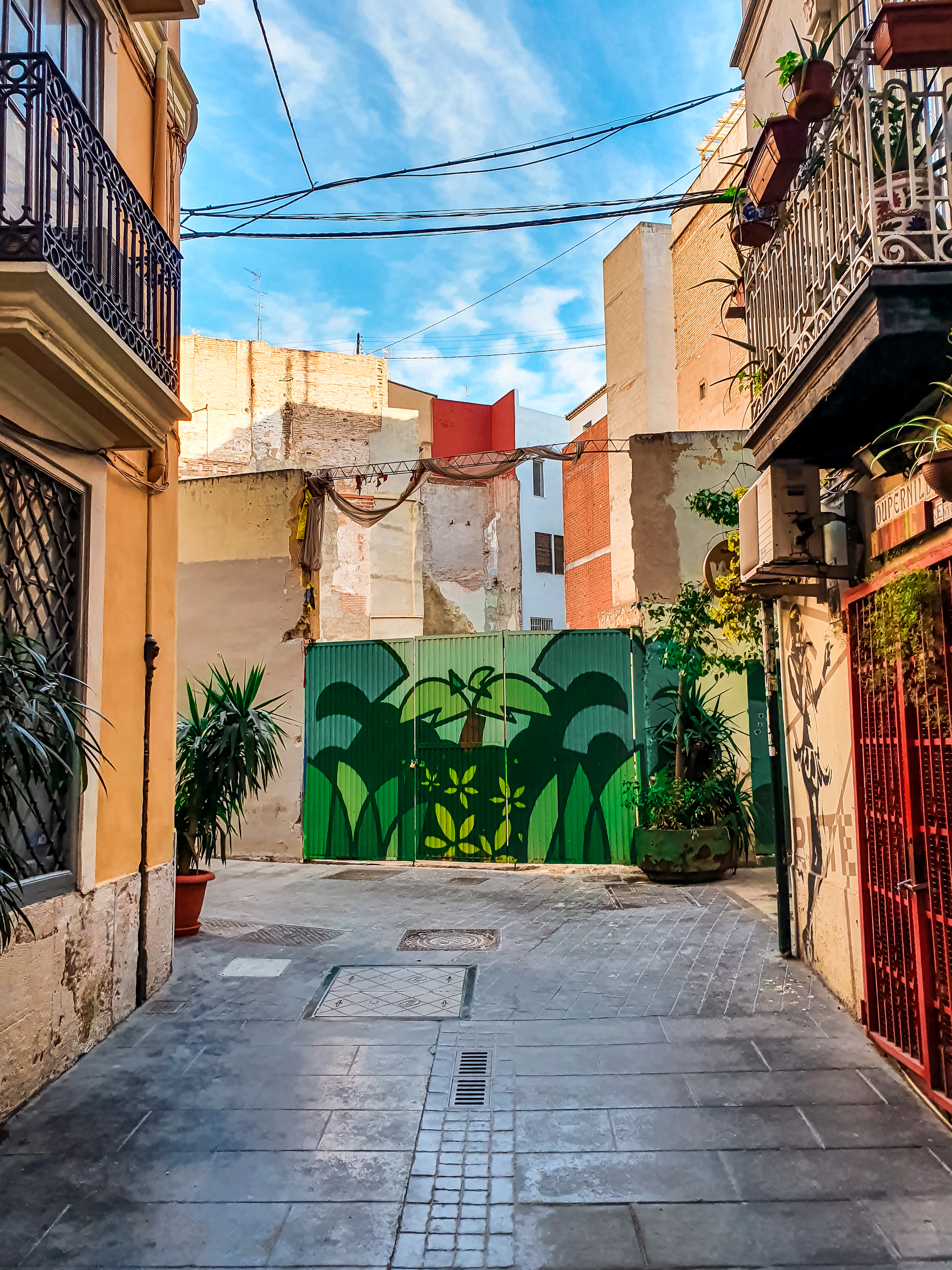 A narrow Valencia alley opens onto a gate painted with a green cactus mural under a blue sky.