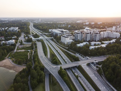 An aerial view of a highway interchange surrounded by greenery and trees. The highway is leading to and through an urban area with multiple buildings, possibly offices or residential apartments. There are cars on the roads and a small beach is visible near a body of water to the left. The sun appears to be setting or rising in the background, casting a warm light over the scene.