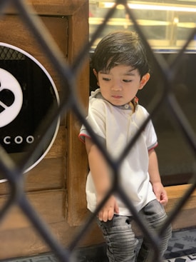 A young child with short black hair is looking downwards while leaning against a wooden surface. The child is wearing a white shirt with red accents and gray pants. The image has a blurred black diagonal grid in the foreground.