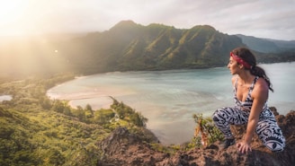 A person wearing patterned workout clothes and a red bandana sits on a rocky ledge overlooking a scenic bay. The landscape features lush green mountains and a sparkling blue ocean, with sunlight streaming in from the left.