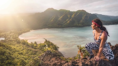 A person wearing patterned workout clothes and a red bandana sits on a rocky ledge overlooking a scenic bay. The landscape features lush green mountains and a sparkling blue ocean, with sunlight streaming in from the left.