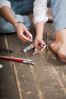 A person sitting on a wooden floor, holding a tube of paint in one hand while squeezing out paint with the other. Several paintbrushes and another tube of paint rest on the floor nearby. The individual is wearing blue jeans and a white shirt, with their bare foot visible in the frame.