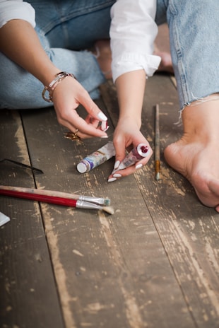 A person sitting on a wooden floor, holding a tube of paint in one hand while squeezing out paint with the other. Several paintbrushes and another tube of paint rest on the floor nearby. The individual is wearing blue jeans and a white shirt, with their bare foot visible in the frame.