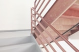 A metal staircase with brown railings and light gray steps is positioned against a plain white wall. The staircase appears to be part of an indoor structure, reflecting minimalistic design elements.