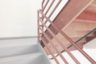 A metal staircase with brown railings and light gray steps is positioned against a plain white wall. The staircase appears to be part of an indoor structure, reflecting minimalistic design elements.