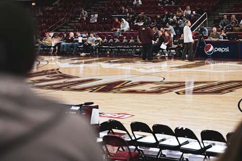 A basketball court with a glossy wooden floor featuring markings and logos. Spectators sitting in rows of maroon seats along the side. A man standing near other people, some are busy with notebooks and electronic equipment. A large Pepsi logo is visible on a sign near the sitting area.