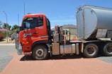 A large red fuel tanker truck is parked on a brick-paved area. The truck has the logo 'Geraldton Fuel' on its side. The tanker is silver and labeled 'Combustible Liquid.' Houses and a street lamp are visible in the background under a clear blue sky.