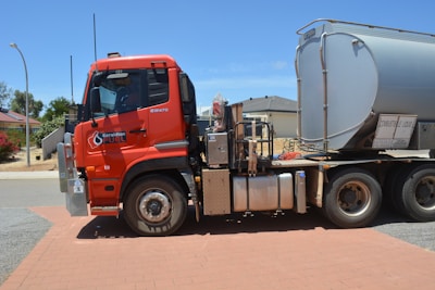 A large red fuel tanker truck is parked on a brick-paved area. The truck has the logo 'Geraldton Fuel' on its side. The tanker is silver and labeled 'Combustible Liquid.' Houses and a street lamp are visible in the background under a clear blue sky.