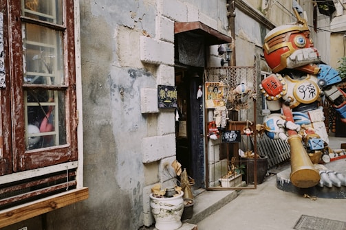 A rustic storefront with an eclectic mix of decor, including a large colorful robot statue on the right. The building has weathered stone walls and a wooden window with peeling paint on the left. Various signs and plants adorn the entryway, adding to the unique charm.