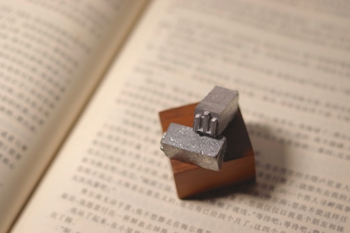 Two metal typesetting blocks rest on a wooden block, placed on an open book with text in an Asian script.