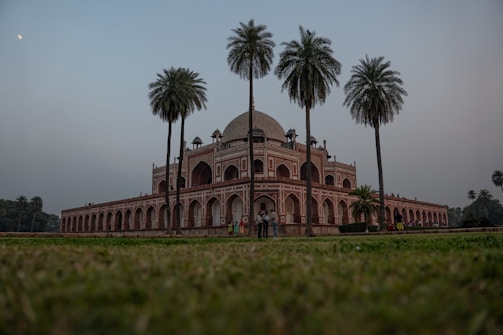 A large, historical building with Mughal architecture featuring a central dome and intricate arches stands amidst a well-maintained garden. Tall palm trees frame the structure, and a few people are gathered around the base of the building. The sky is overcast with a slight view of the moon.