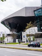 Futuristic architecture of a building with reflective glass elements, surrounded by trees and a street. The foreground features a parked Mini Cooper car, and a BMW Group sign is visible near the entrance. Two people are standing near the sign, and the scene is complemented by three BMW flags.