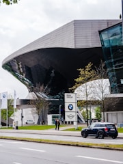 Futuristic architecture of a building with reflective glass elements, surrounded by trees and a street. The foreground features a parked Mini Cooper car, and a BMW Group sign is visible near the entrance. Two people are standing near the sign, and the scene is complemented by three BMW flags.