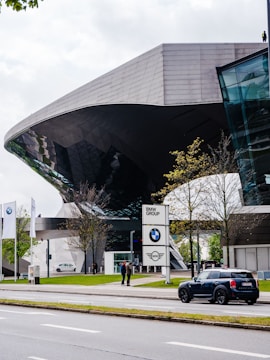 Futuristic architecture of a building with reflective glass elements, surrounded by trees and a street. The foreground features a parked Mini Cooper car, and a BMW Group sign is visible near the entrance. Two people are standing near the sign, and the scene is complemented by three BMW flags.