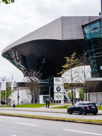 Futuristic architecture of a building with reflective glass elements, surrounded by trees and a street. The foreground features a parked Mini Cooper car, and a BMW Group sign is visible near the entrance. Two people are standing near the sign, and the scene is complemented by three BMW flags.