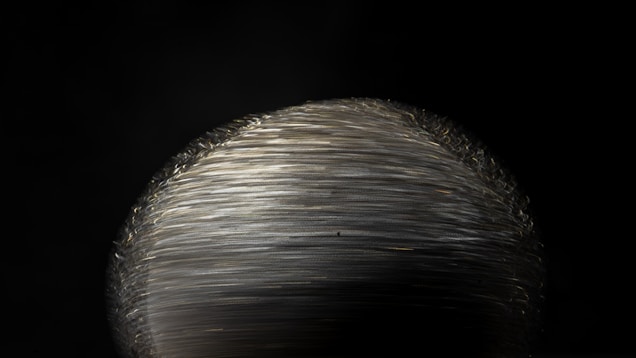 A close-up view of a metallic, shiny object with intricate texture and layered strands. It resembles a spool of tightly wound wire or thread, predominantly silver in color, standing out against a dark background.