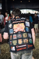 A person is wearing a heavily patched denim jacket featuring various colorful logos and names of rock and metal bands. The scene is set in a casual, crowded environment with several people blurred in the background. The patches include names like Sabbath Bloody Sabbath, Metallica, and System of a Down.
