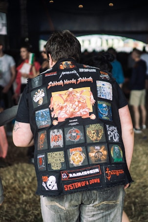 A person is wearing a heavily patched denim jacket featuring various colorful logos and names of rock and metal bands. The scene is set in a casual, crowded environment with several people blurred in the background. The patches include names like Sabbath Bloody Sabbath, Metallica, and System of a Down.