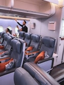 Airplane seating area with several rows of well-cushioned seats in a neutral color, accented by orange pillows. Overhead compartments are visible, with one open. Passengers in the background are either seated or standing, with one individual stowing items in the overhead bin. The ambient lighting gives a calm atmosphere to the cabin.
