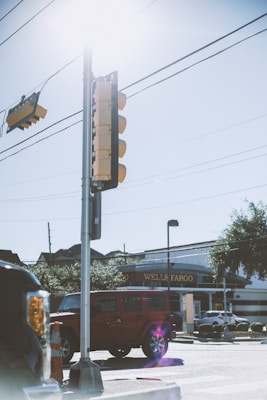 A sunny street scene with a traffic light in the foreground, a red vehicle waiting at a crosswalk, and a Wells Fargo bank branch in the background. A construction cone is placed near the vehicle, and power lines run across the sky.
