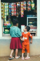 Two children stand in front of a small shop with various colorful snack packets hanging outside. The girl is wearing a green top and a red skirt, while the boy wears an orange shirt and shorts. The shop is filled with snacks and goods.