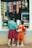 Two children stand in front of a small shop with various colorful snack packets hanging outside. The girl is wearing a green top and a red skirt, while the boy wears an orange shirt and shorts. The shop is filled with snacks and goods.
