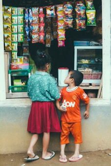 Two children stand in front of a small shop with various colorful snack packets hanging outside. The girl is wearing a green top and a red skirt, while the boy wears an orange shirt and shorts. The shop is filled with snacks and goods.